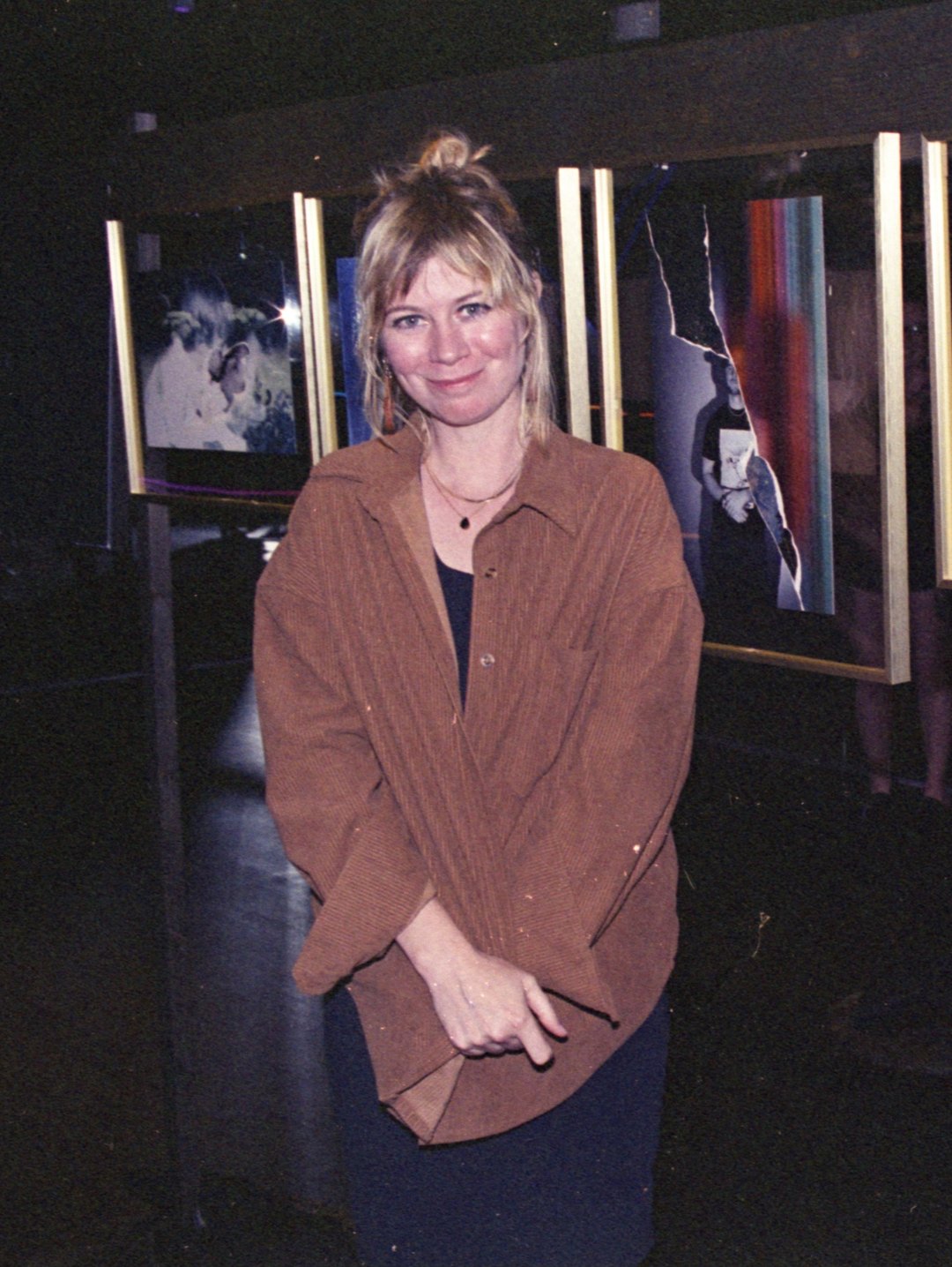 Kate in a brown corduroy shirt, smiling in a dark gallery in front of framed photographs.