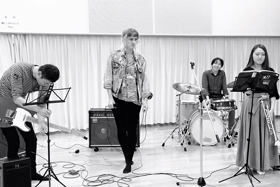 Black-and-white photo of a singer performing with a guitarist, a drummer, and a trumpet player in a rehearsal hall.