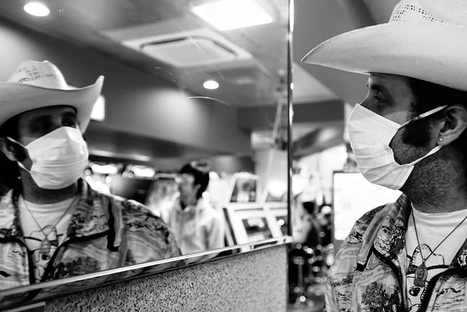 Black-and-white photo of two musicians in cowboy hats and face masks, one reflected in a window.