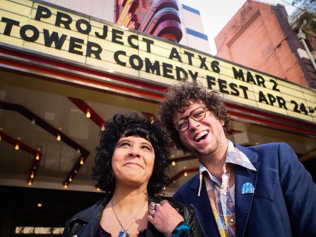 Two musicians grinning beneath a theater marquee that reads “Project ATX6” and “Tower Comedy Fest.”