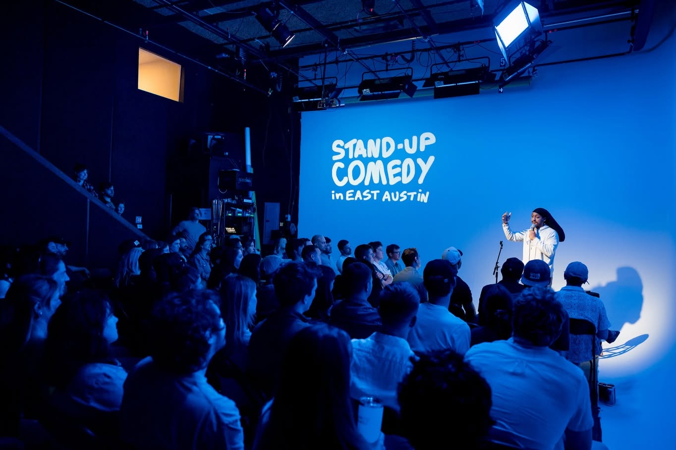 A comedian performing under a spotlight to a packed audience, in front of a blue wall with “Stand-Up Comedy in East Austin” projected on it.