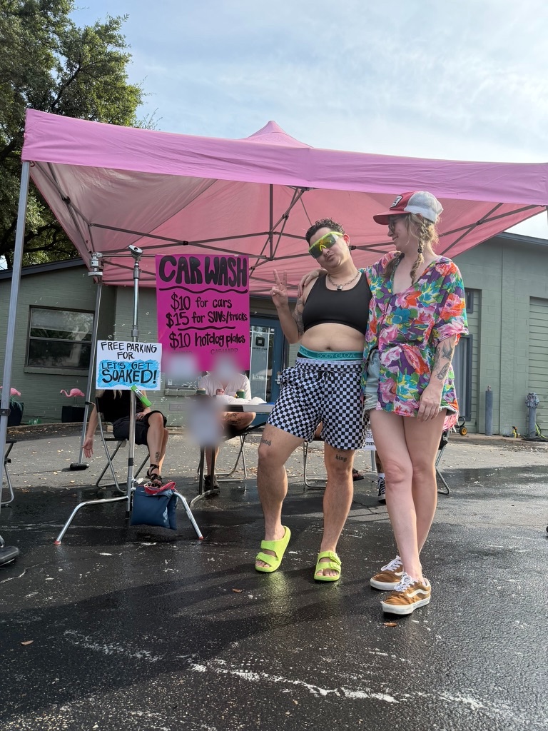 Two people posing in a wet parking lot under a pink canopy, beside a hand-painted car wash price sign and a table where volunteers sit.