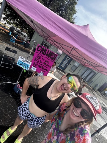 A sunny selfie of two volunteers in sunglasses and swimwear under a pink canopy with a hand-painted car wash sign.