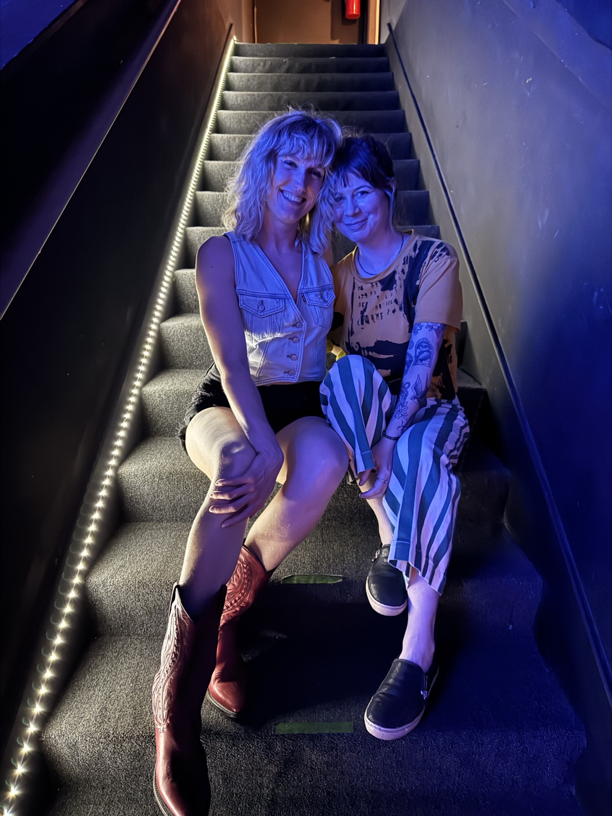 Kate and another person sitting close together and smiling on a carpeted staircase lit blue, with a strip of small lights along the wall.