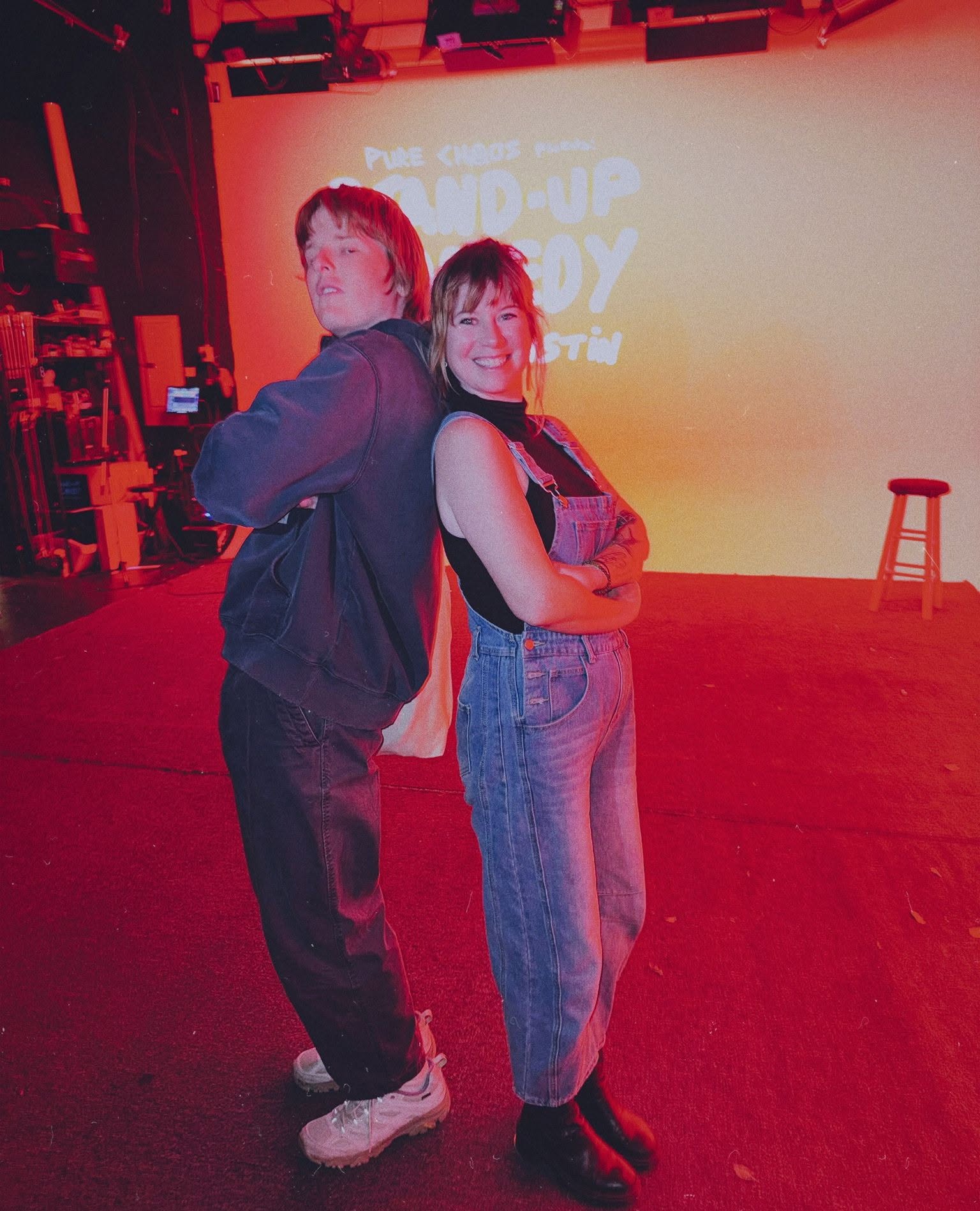 Kate and another person posing back to back with arms crossed on a red-lit stage, with a projected stand-up comedy sign and a stool behind them.