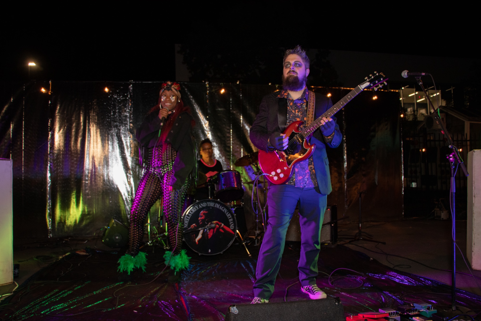 Noella Grey and the Imaginary Band performing at night in front of a silver foil backdrop, with a singer, a drummer and a guitarist.