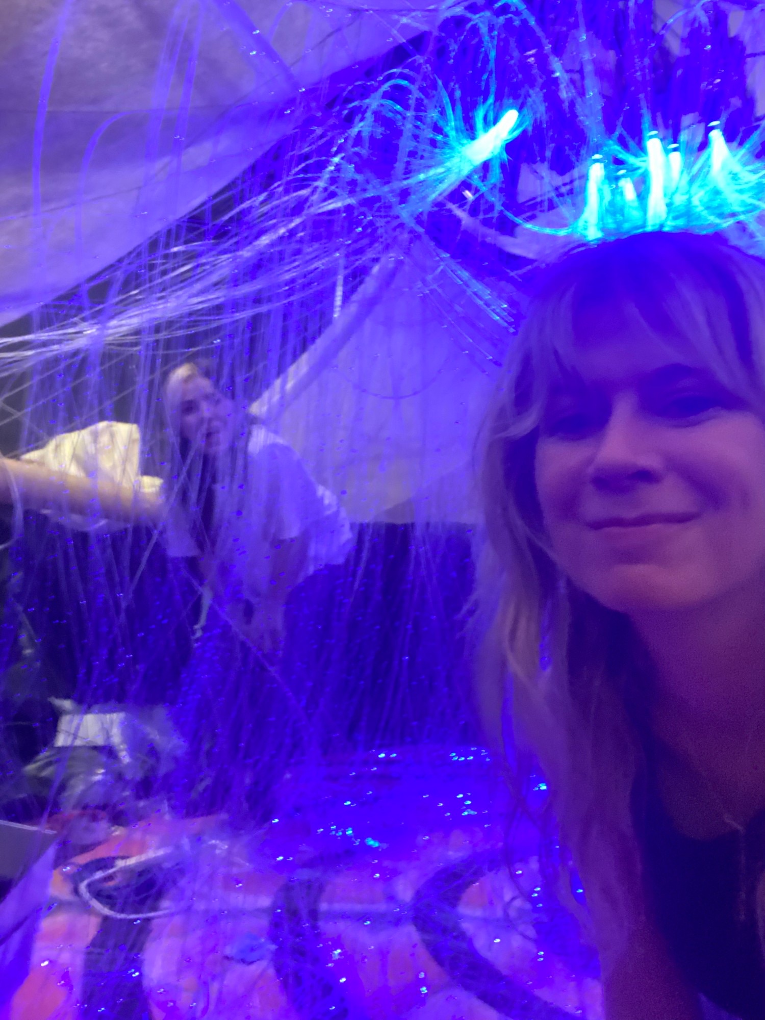 Kate smiling in a room glowing blue and purple, surrounded by hanging strands of fiber-optic light, with another person behind her.