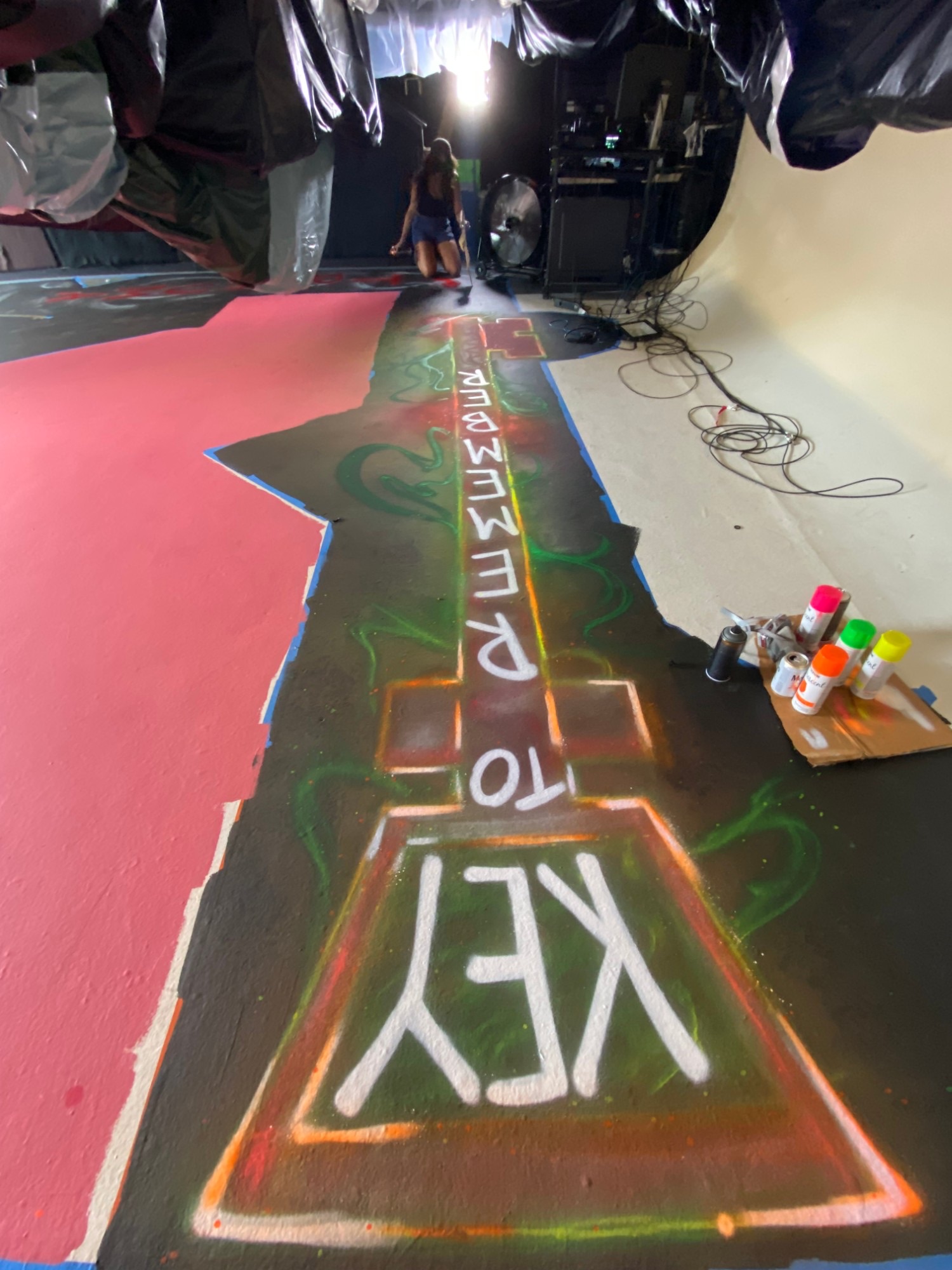 During the build, a giant key spray-painted in neon on the studio floor, with cans of paint beside it and an artist kneeling at the far end.