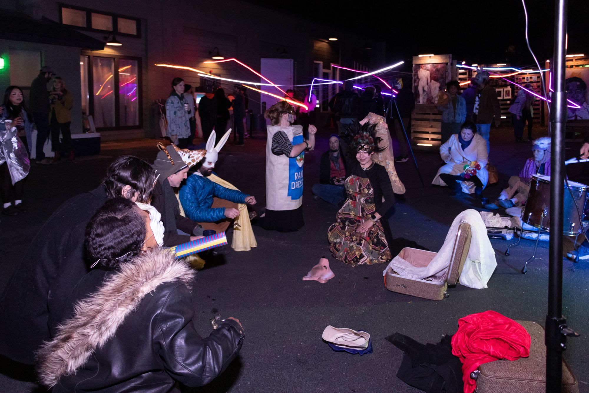 At night under the light installation, people in masks and costumes, including a bunny mask, a giraffe hat and a ranch dressing bottle, sit in a circle with suitcases of props.