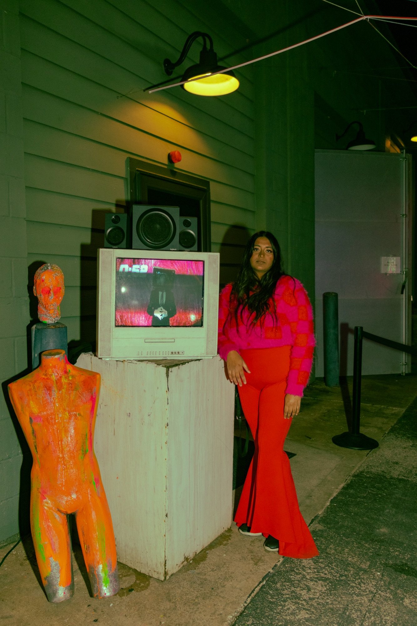 An installation outside a building: an old TV playing video on a white pedestal with speakers on top, beside a paint-covered orange mannequin, with a person in red standing next to it.