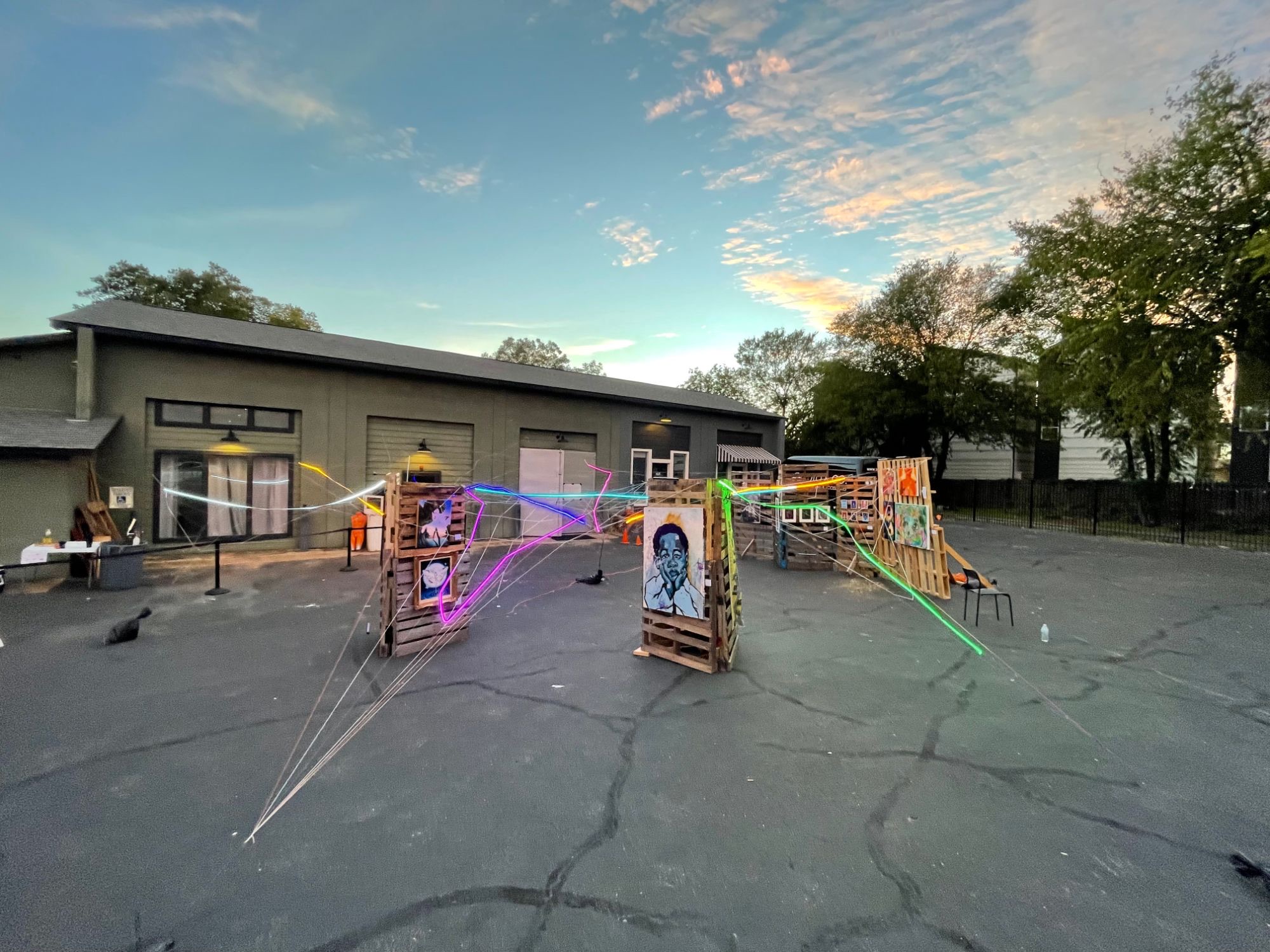At sunset outside the studio, colored light strands and string stretch between pallet towers hung with paintings, including a large portrait of a child resting his face in his hands.