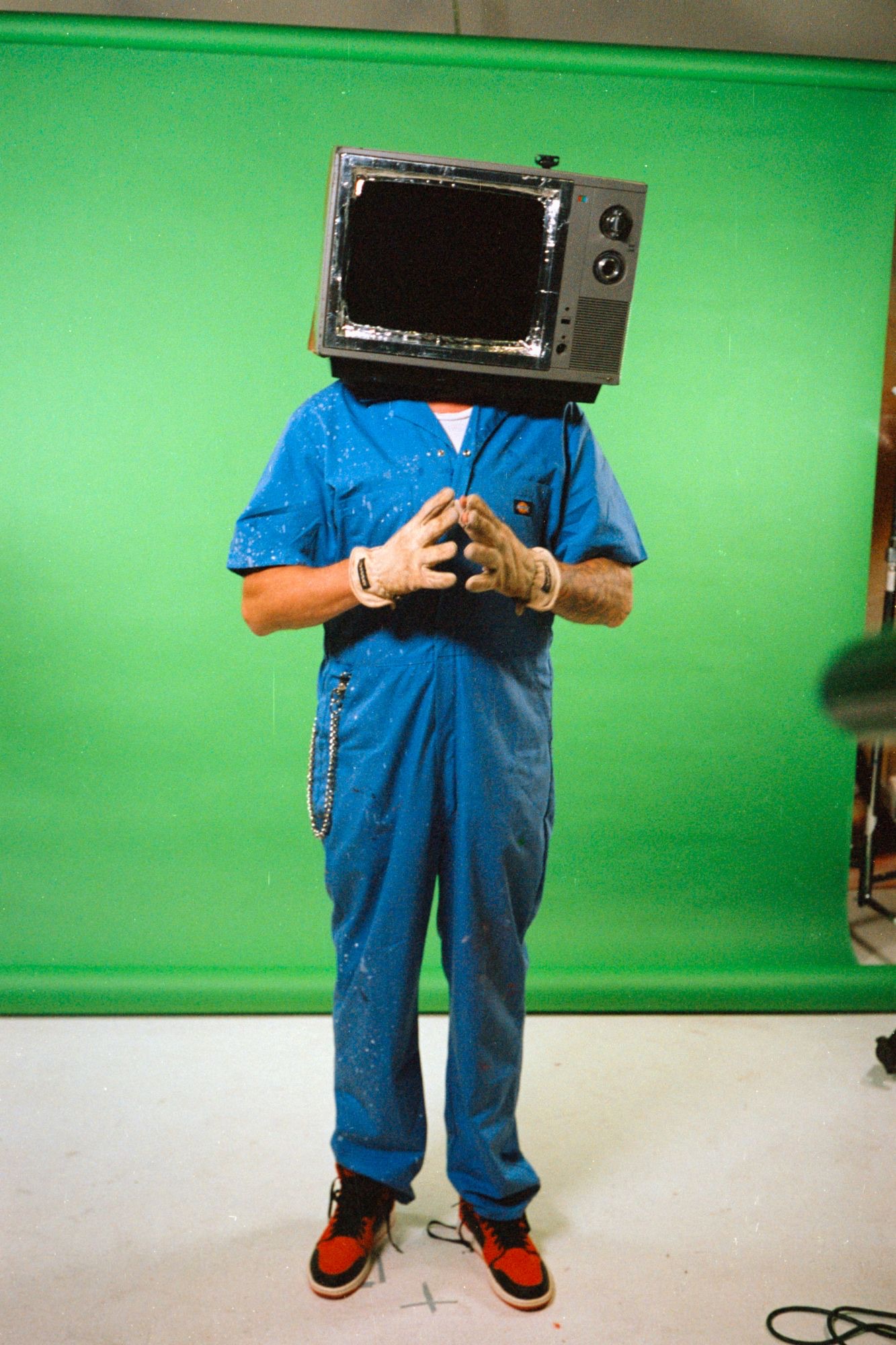 TVHead stands in front of a green screen in blue coveralls, work gloves and red sneakers, with an old television over his head.