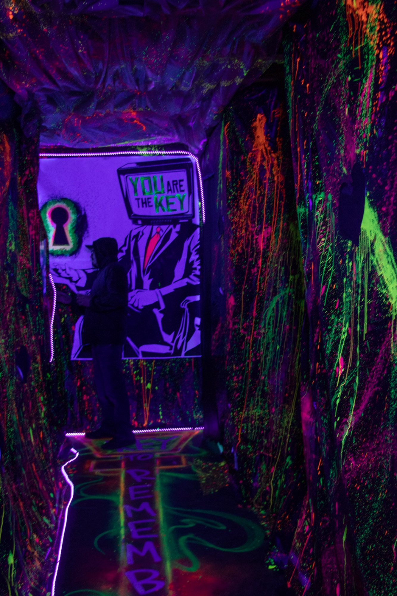 A person stands in a narrow corridor covered in glowing neon paint splatters, facing a lit mural of a figure with a TV for a head and the words “You are the key.”