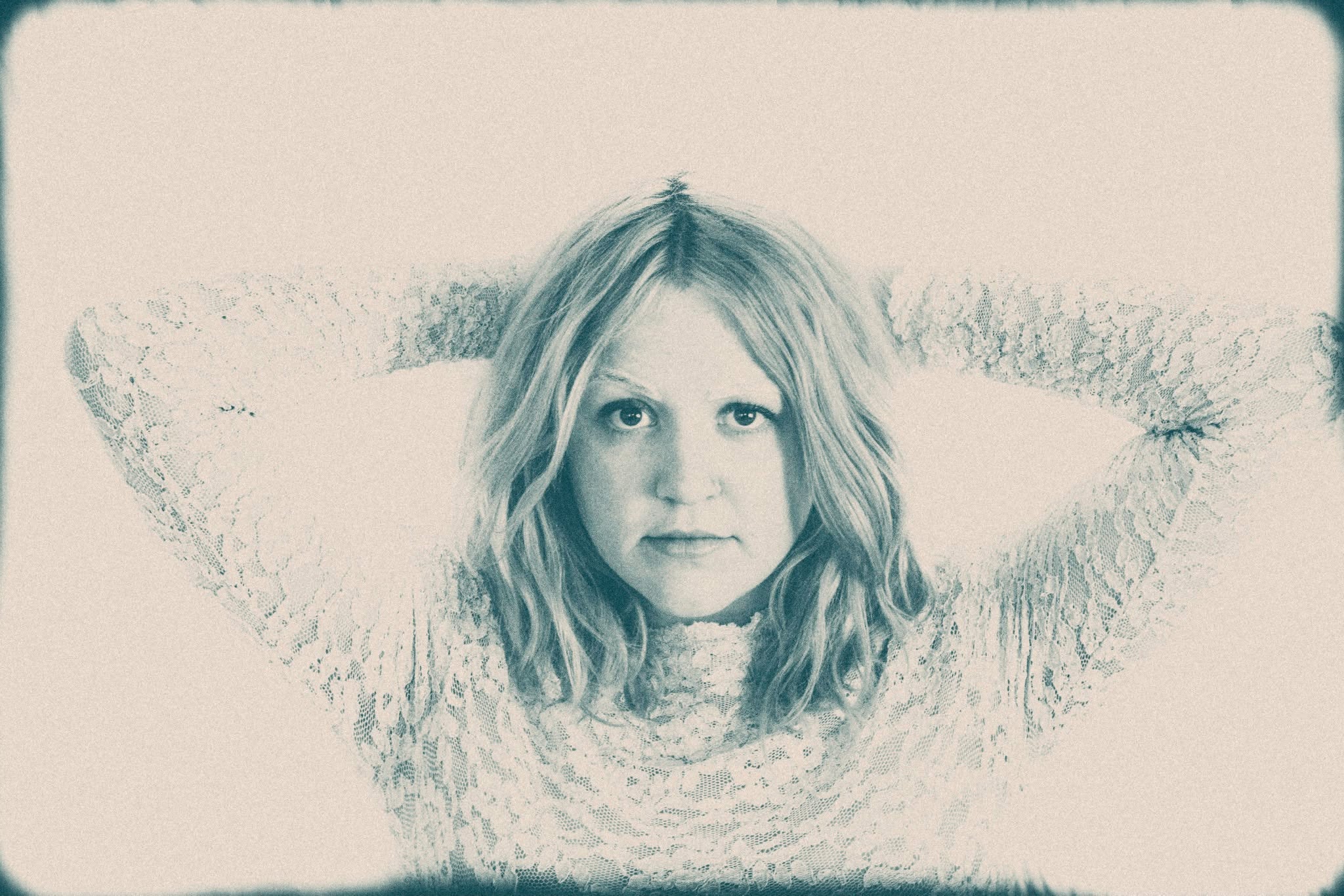 Soft monochrome portrait of Carrie Fussell with her arms raised behind her head, framed by a pale vignette.