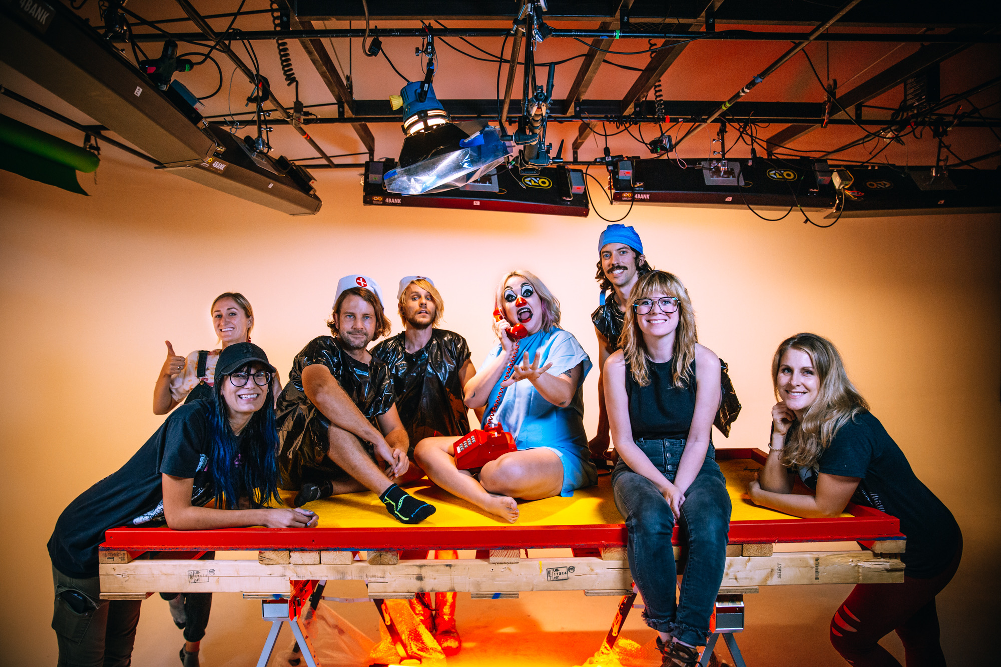 Cast and crew pose together on a raised yellow platform in the studio, one in clown makeup holding a red telephone.