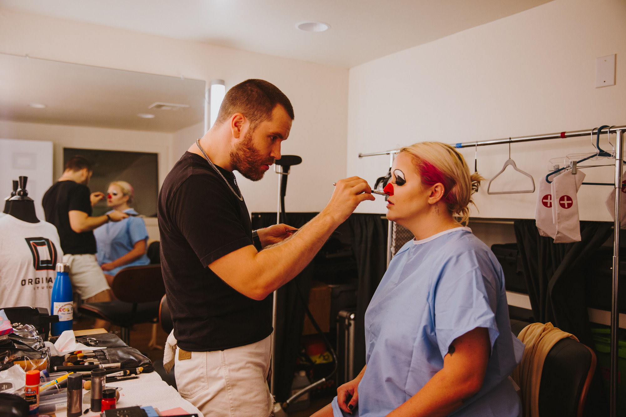 In a dressing room, a makeup artist paints a red clown nose on a performer in a hospital gown.