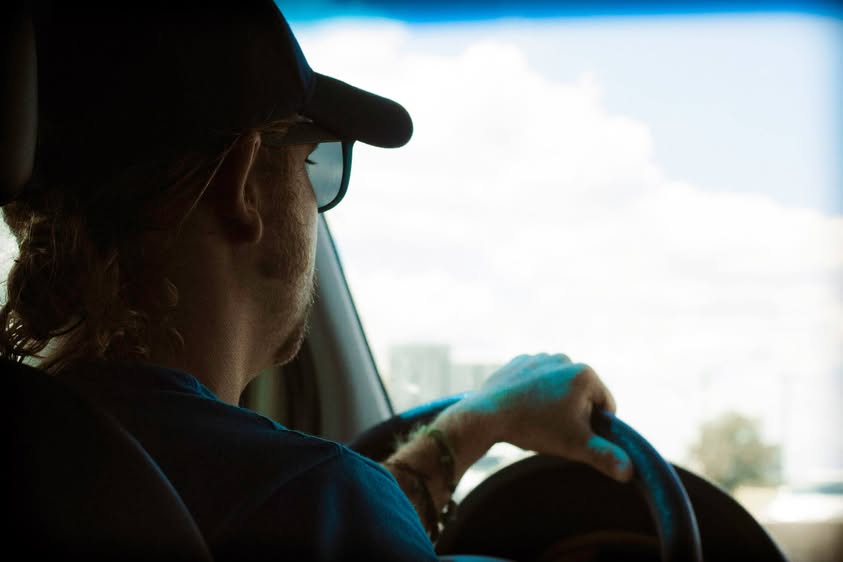 A driver in a cap and sunglasses at the wheel, with bright sky through the windshield.