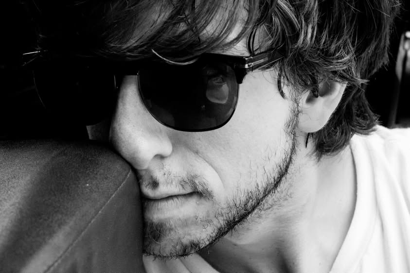 Black-and-white portrait of a musician in dark sunglasses resting his head against a seat.